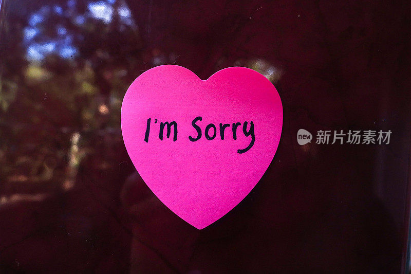 “对不起”这个词写在一张粉红色的便利贴上，在红色的背景上形成一个心形。用便条道歉的概念