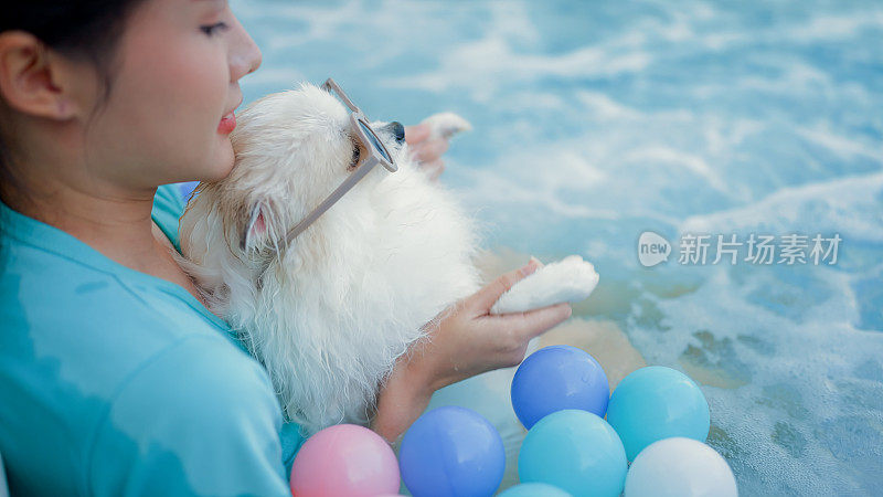 泳池边，一名亚洲年轻女子和她的白色博美犬站在充气玩具火烈鸟上。夏天，户外的乐趣和生活方式