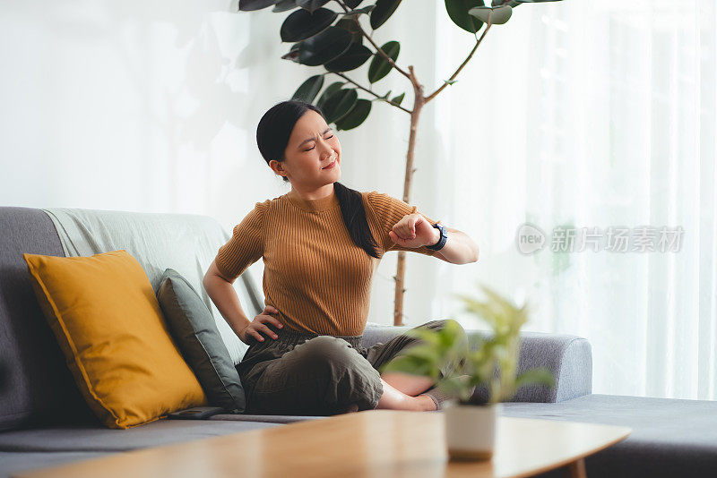 一名亚洲妇女坐在家里客厅的沙发上，饱受背痛之苦。