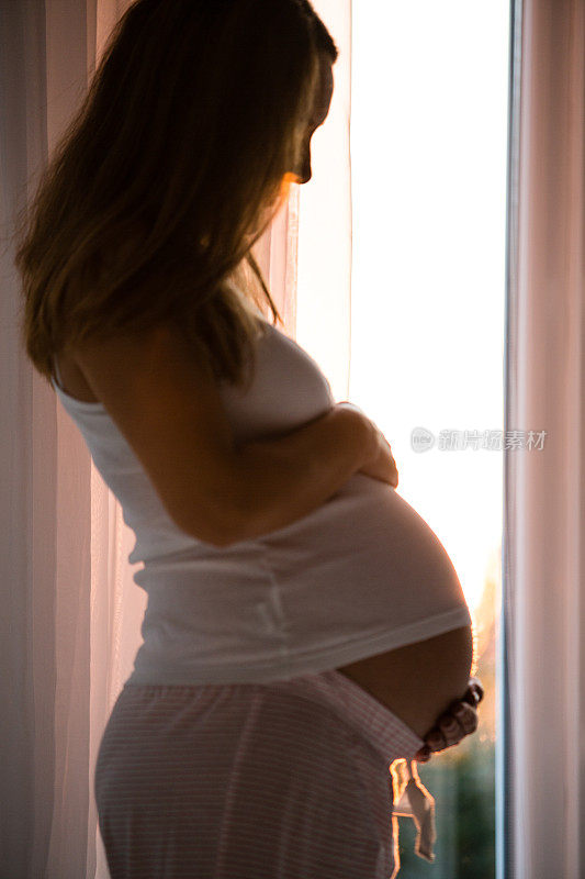 一个孕妇抱着她的肚子站在窗边