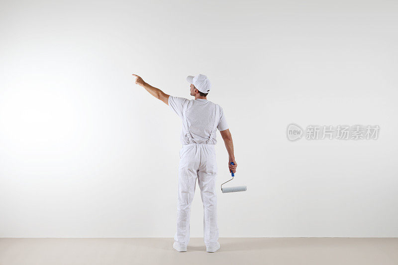 后视图画家用手指指着空白的墙壁，用油漆滚筒，隔离在白色的房间