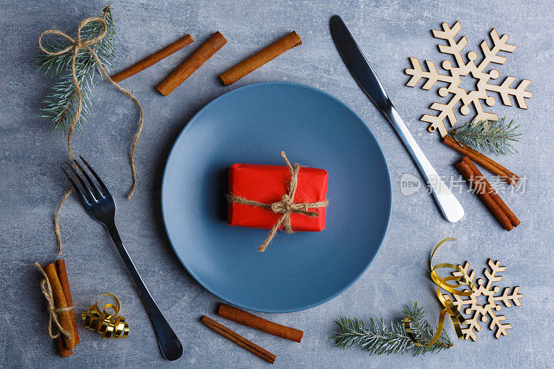 一个蓝色盘子和一个小礼品盒旁边的刀叉，肉桂棒，圣诞树和雪花。