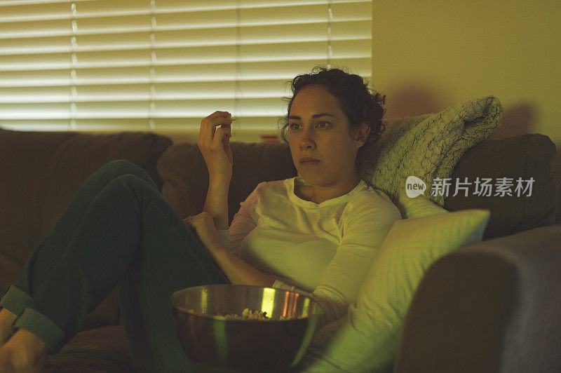 一名20多岁的年轻女子抱着一碗爆米花依偎在沙发上。