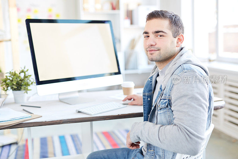 微笑英俊的年轻男性搜索引擎优化专家胡茬坐在办公室的椅子上，看着相机，而工作与电脑在办公室
