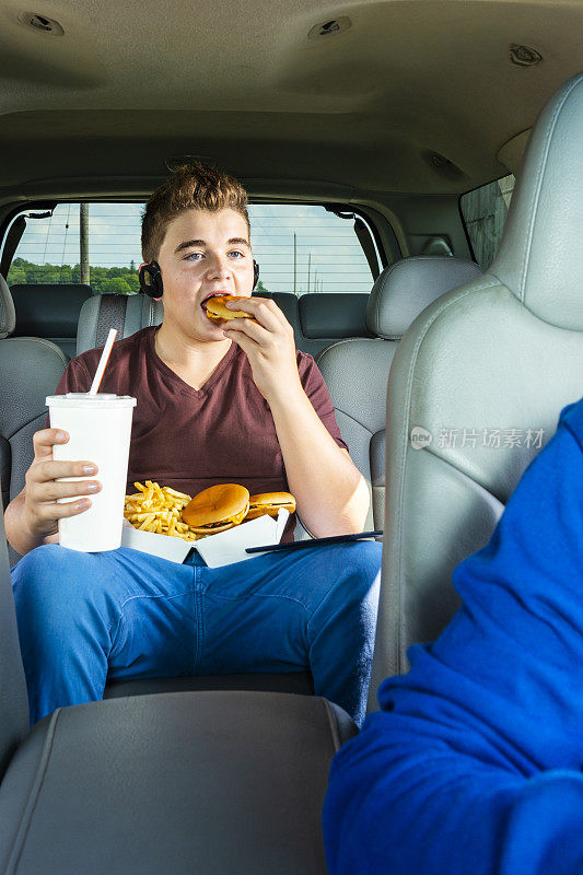 一个十几岁的男孩在汽车后座上吃着外卖的汉堡和薯条