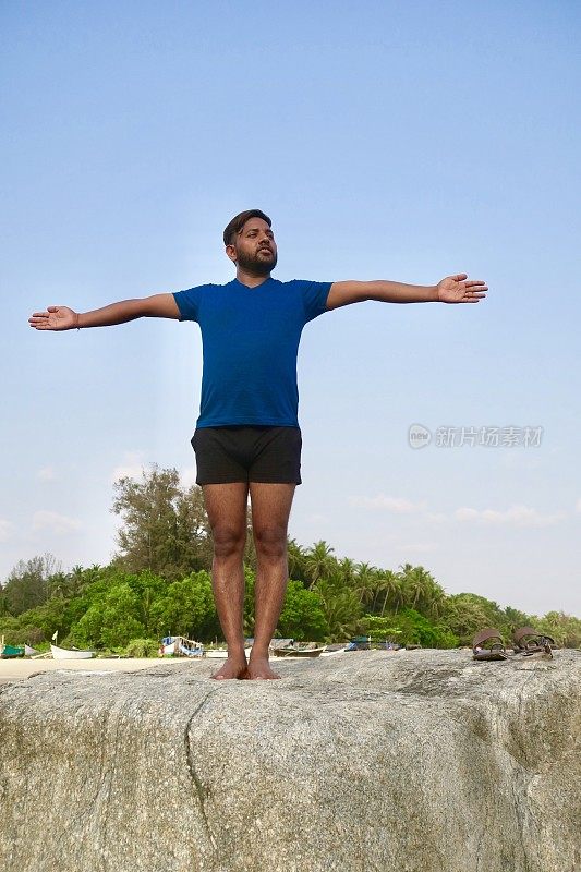 印度男子练习山姿势伸出手(山式伸出手)瑜伽姿势在岩石，水的边缘，帕洛伦海滩，果阿，印度