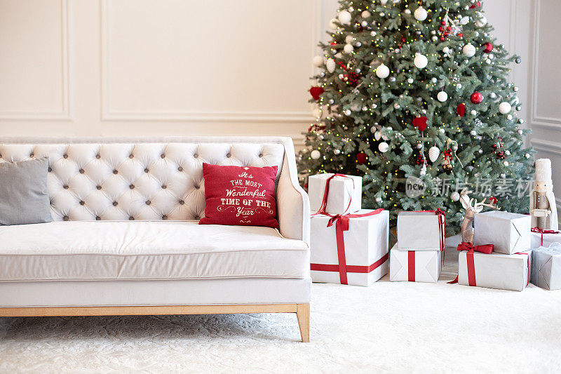 豪华客厅内饰以沙发装饰别致的圣诞树、礼物和枕头。红色色调的经典内饰。圣诞节在家里。圣诞树背景上的米色沙发。