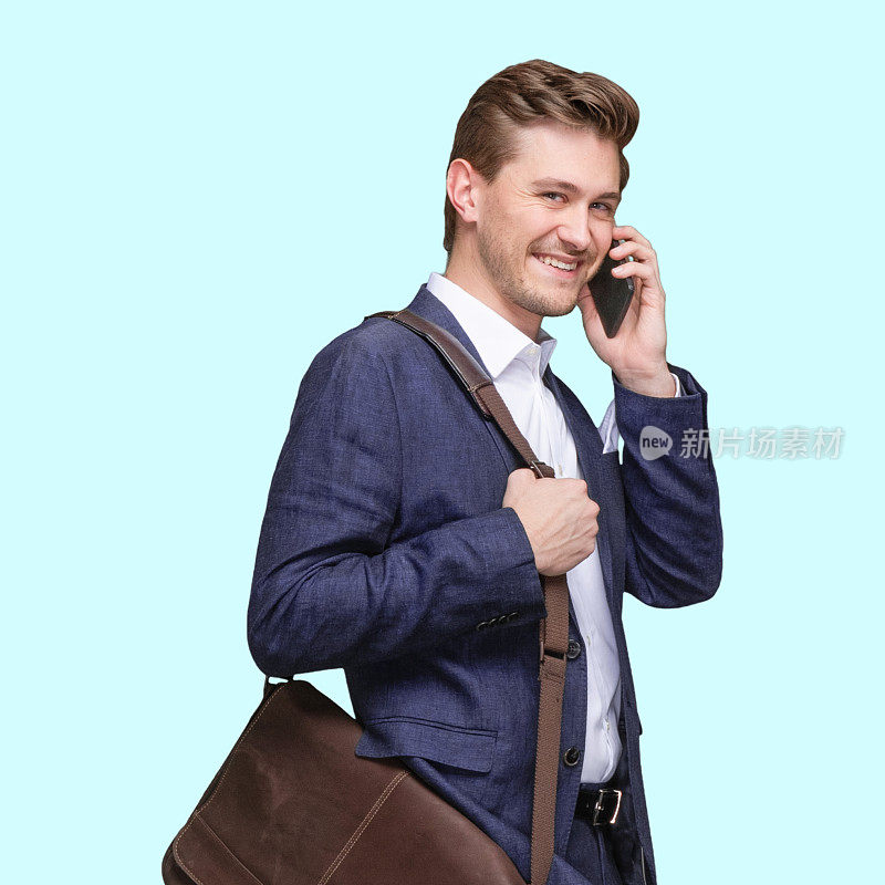 白人年轻男性商务人士站在蓝色背景前，肩挎包，手持包，使用手机