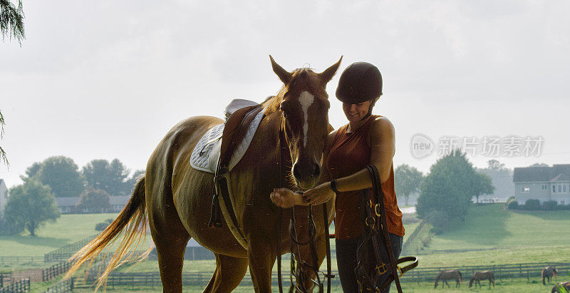 一个三十多岁的年轻女人戴着头盔调整她的棕色马的笼头在一个阳光明媚的日子里，在一个有农舍，牧场，和放牧的马在背景