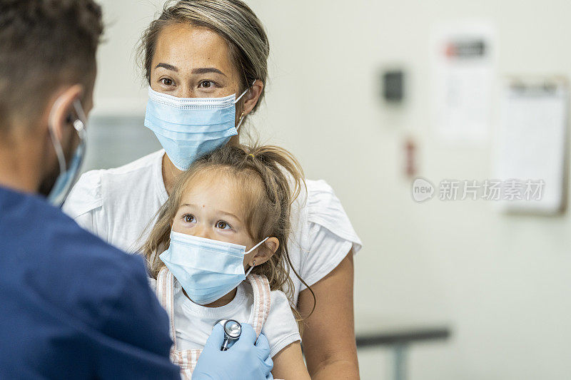 儿科医生正在检查一位戴着口罩坐在母亲膝盖上的年轻女性病人。
