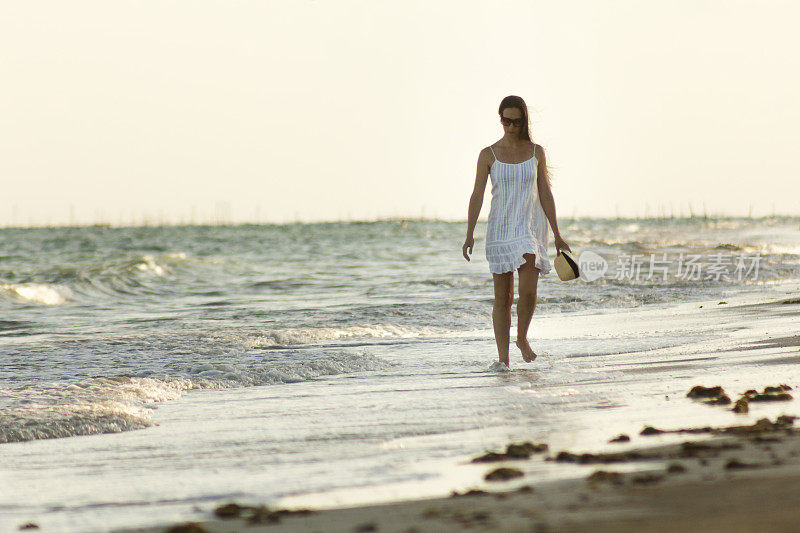 在巴西东北部的阿拉格斯，美丽的女人走在日落的海滩上的肖像。她穿着白色的衣服，戴着太阳镜和草帽