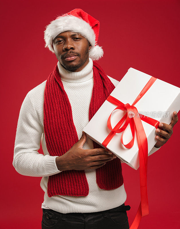 一个皮肤黝黑的男人，穿着毛衣，戴着圣诞帽，拿着一个礼品盒，愤怒地皱着眉头，撅着嘴