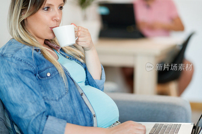 孕妇坐在家里的沙发上，拿着笔记本电脑喝着咖啡
