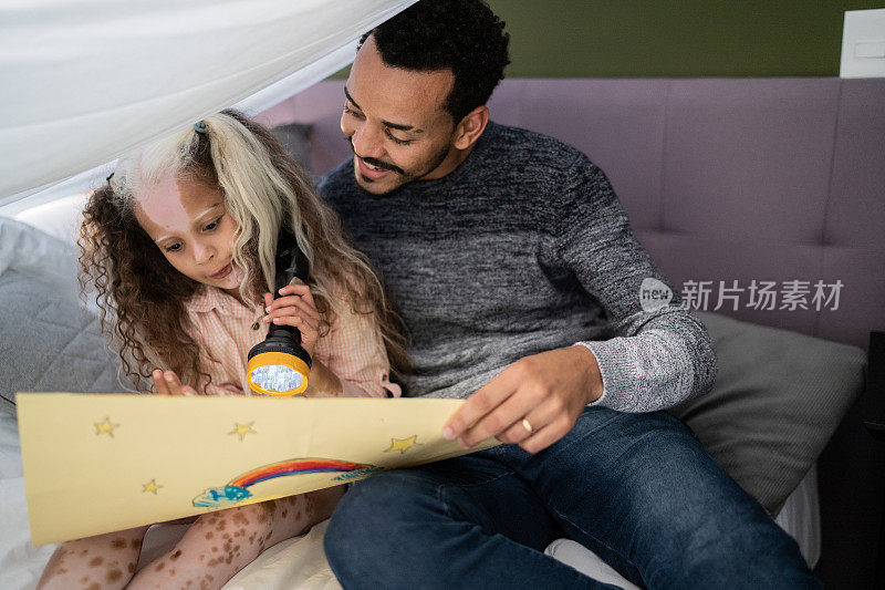 父亲和女儿在家里盖着毯子的帐篷里玩耍或读书——女孩有花语