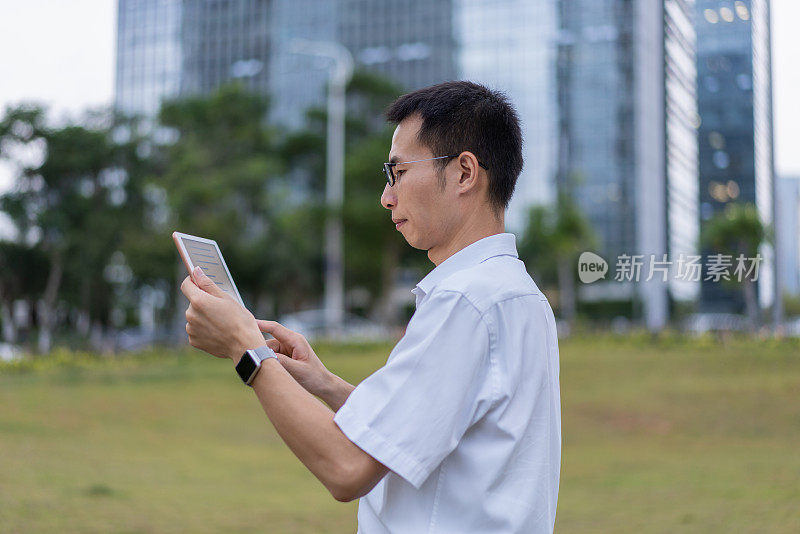 一位亚洲男性白领正在办公大楼外的草地上使用平板电脑