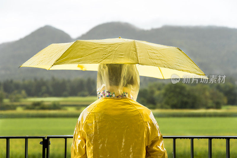 从公寓阳台看雨天的黄色雨衣和雨伞的女孩的后视图