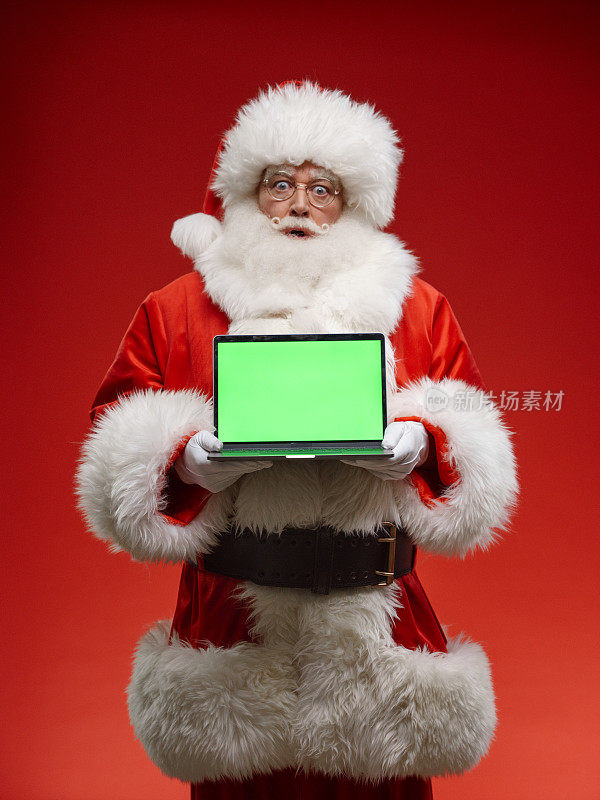惊讶的圣诞老人在一台笔记本电脑的屏幕上展示了chroma键，他站在一个渐变的红色背景上，在一个专业工作室的灯光下