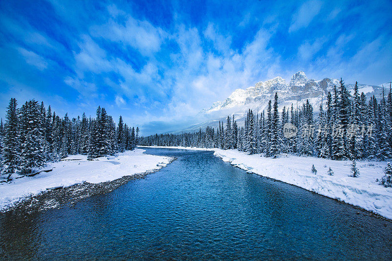 田园诗般的冬季景色，明亮的蓝色融雪河，积雪覆盖的山脉景观和明亮的蓝色天空与戏剧性的云的形成