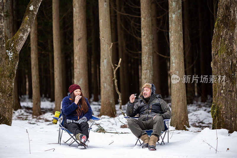 一对夫妇在雪林风景中放松地坐在露营椅上吃着饭团