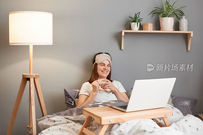 微笑快乐满足的女人戴着笔记本电脑的睡眠面具，躺在床上，喝着热的美味的咖啡，看电影，笑得很开心，享受慵懒的早晨。