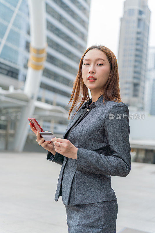 一名年轻的亚洲女商人穿着灰色的办公套装，在商业区用她的智能手机使用信用卡进行在线交易，她微笑着自信