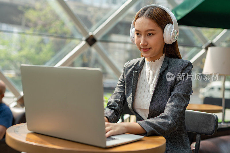混合式工作风格。年轻的亚洲女性成年人与笔记本电脑在咖啡馆工作。戴白色耳机的女商人