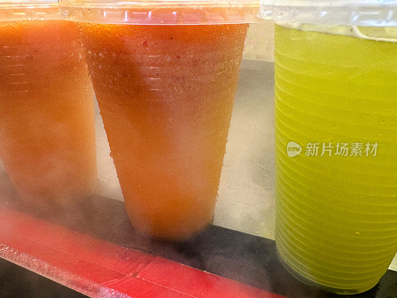 全画幅图像，橙色和柠檬冰沙饮料在有盖的塑料杯，雪筒冰饮料在一次性纸杯覆盖凝结，人工彩色冰沙在金属柜台，重点在前景