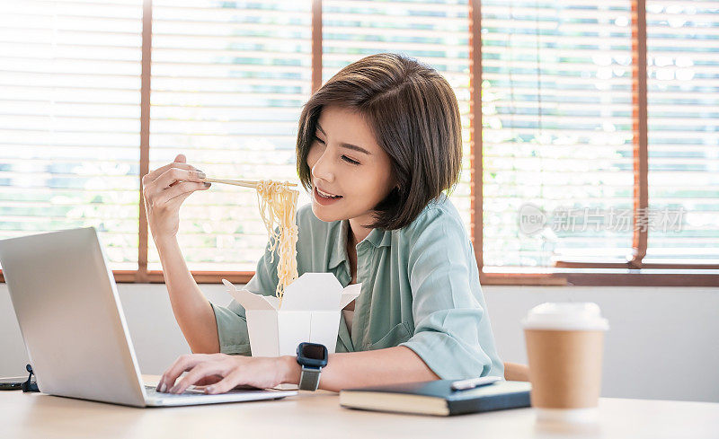 年轻的亚洲女性在办公室用笔记本电脑享受中餐的肖像，在家工作，女员工拿着筷子在工作场所吃外卖面盒饭。