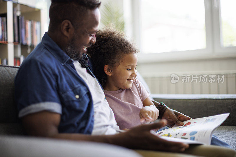 快乐的非裔美国父亲和可爱的混血女儿在家里一起读书，面带微笑。儿童教育发展理念。