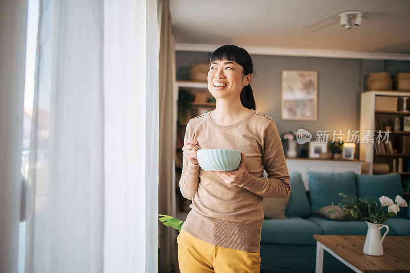 日本中年妇女站在窗后，端着盛有食物的碗。