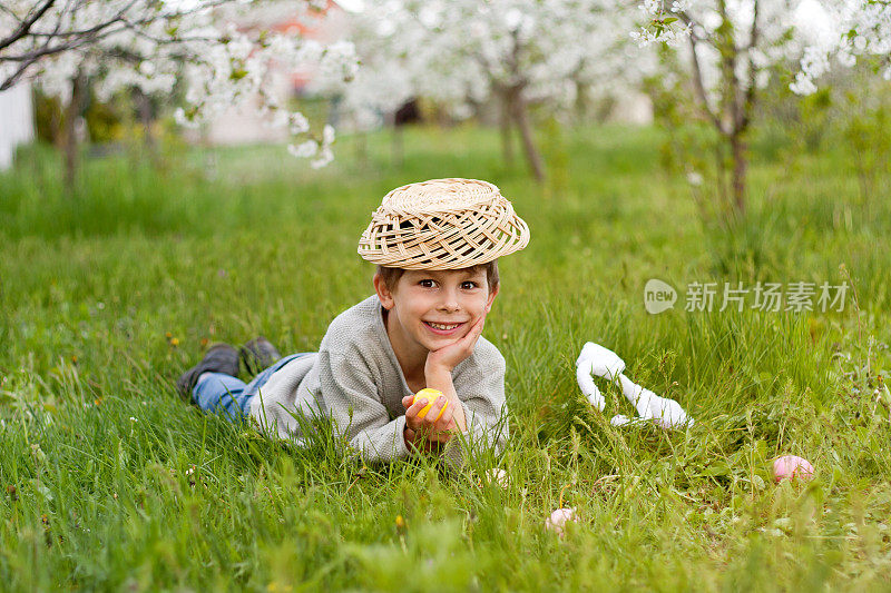 寻找复活节彩蛋。可爱的笑着的男孩，头上顶着篮子，躺在绿色的草地上，篮子里放着复活节彩蛋。复活节