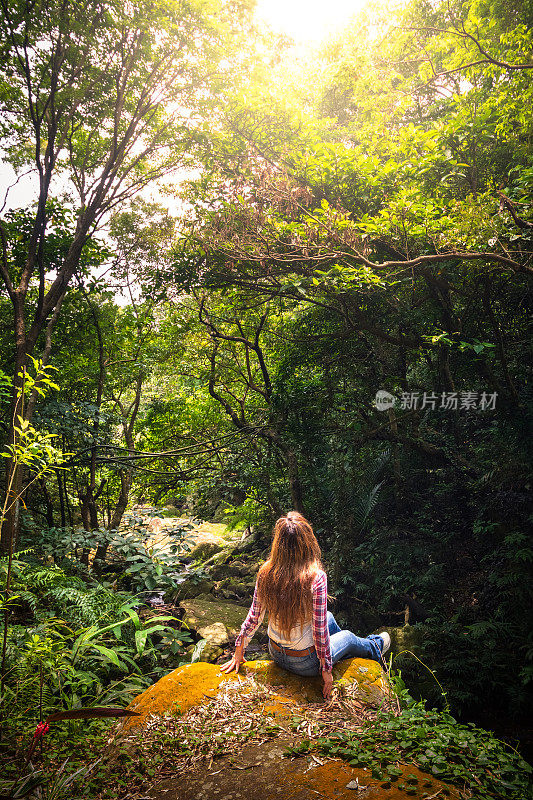 在台湾台北，一名妇女坐在长满青苔的岩石上，小溪沿着元觉瀑布徒步旅行