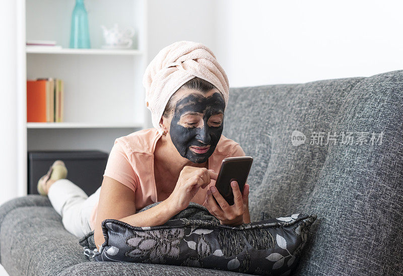 年长妇女带着面膜躺在沙发上，在家里使用手机