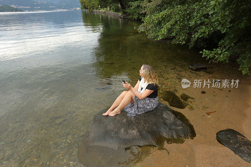 成熟的女人坐在淹没在水里的岩石上发短信