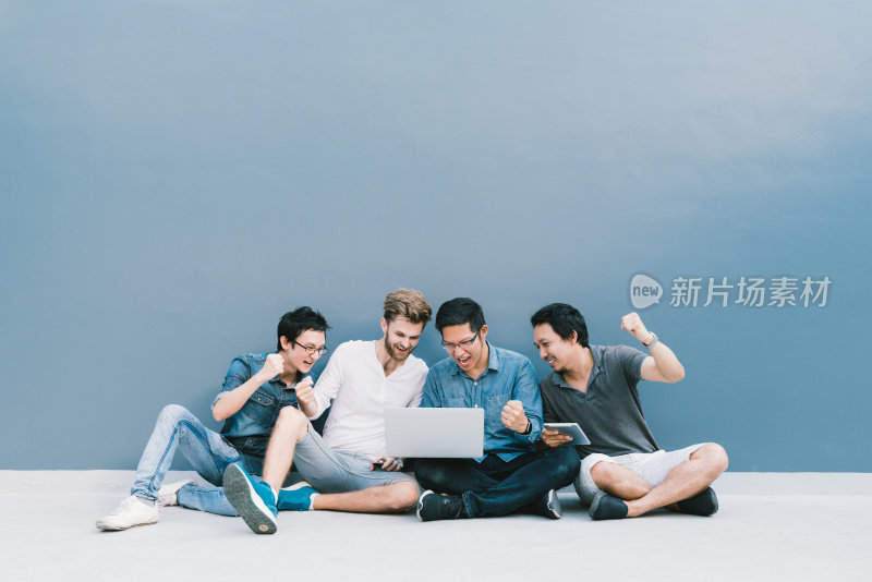 多民族4名男子一起用笔记本电脑庆祝，坐在蓝色墙壁与复印空间。大学生的生活方式，信息技术工具，教育，社交网络，或成功的概念