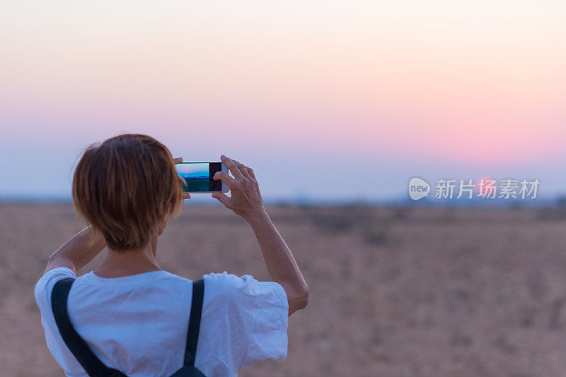 沙漠日落时，女人用智能手机拍照。游客拍摄纳米布沙漠，日落浪漫的天空，旅游目的地纳米比亚，非洲。人们旅行旅游热。