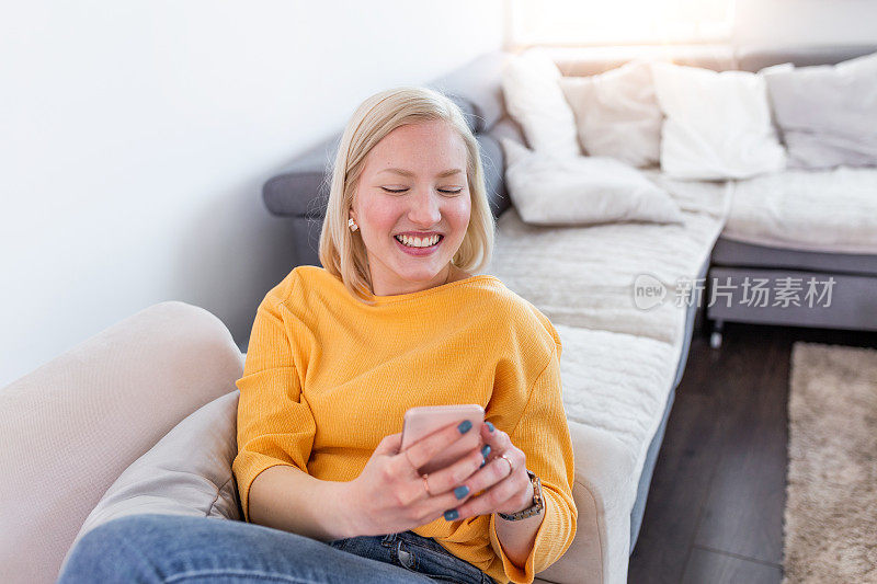 一个微笑的女人坐在家里的沙发上看手机短信的完整镜头。快乐的女孩坐在家里的沙发上看智能手机短信
