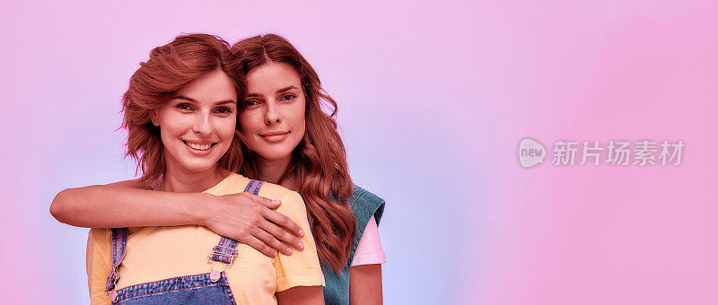 两个迷人的年轻女孩的肖像，双胞胎姐妹对着相机微笑，在粉红色的背景下一起摆出孤立的姿势
