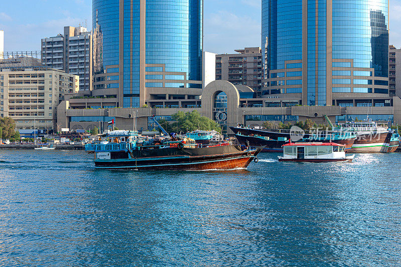 阿拉伯联合酋长国迪拜——一艘满载传统阿拉伯单桅帆船在晨曦中驶入迪拜河;在背景中，德拉一侧的单桅帆船互相停靠;在遥远的背景，办公室和住宅建筑