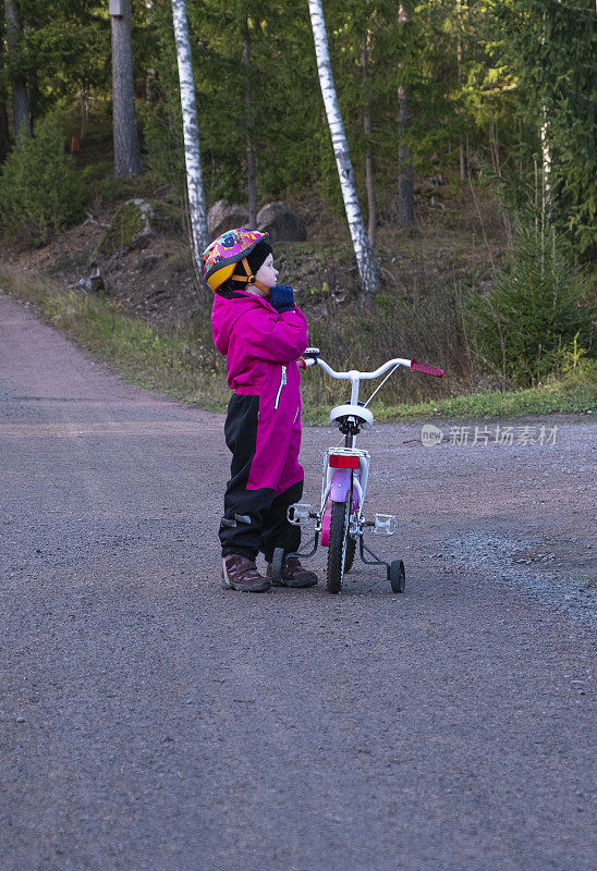 一个5岁的白人女孩，在乡下骑自行车。站在自行车旁边，戴上安全帽。
