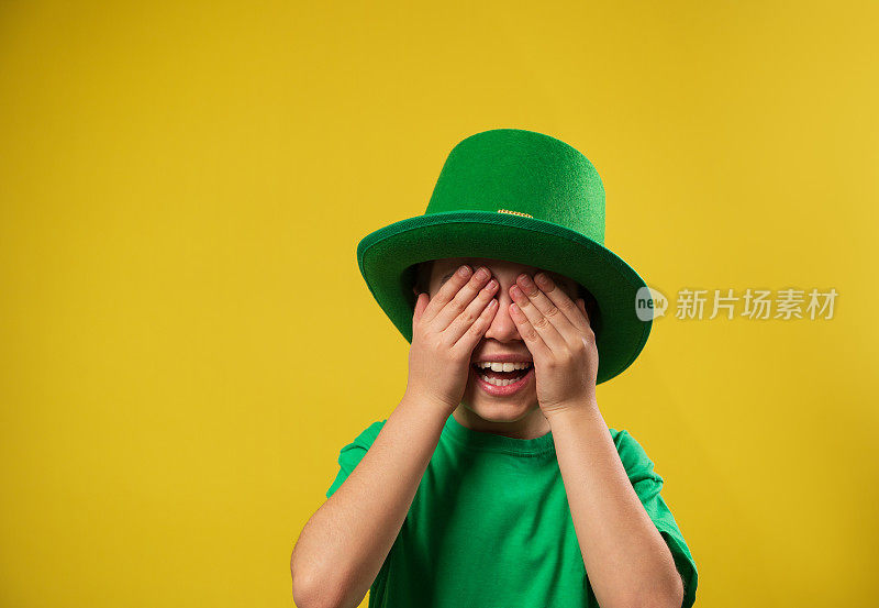 小男孩笑着戴着绿色的爱尔兰妖精帽子，用手掌遮住了眼睛。圣帕特里克节在黄色背景和复制空间