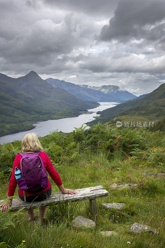 一位成年女性背包客在苏格兰Kinlochleven的山上徒步旅行时，坐在长凳上欣赏利文湖的景色。