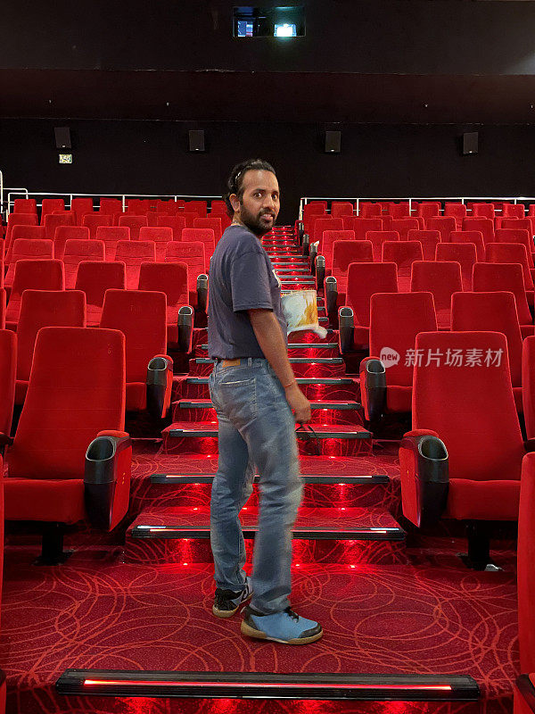 印度男子拿着一桶爆米花爬上电影院的台阶，一排排的红色座位上有饮料架，靠过道的座位上有台阶，看着镜头