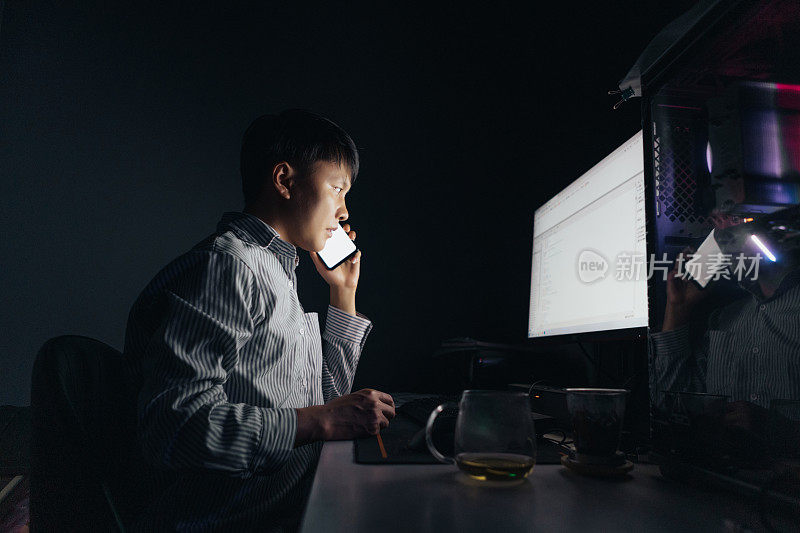 一名亚洲男子晚上在电脑前加班，用智能手机开视频会议，这种生活方式很不健康