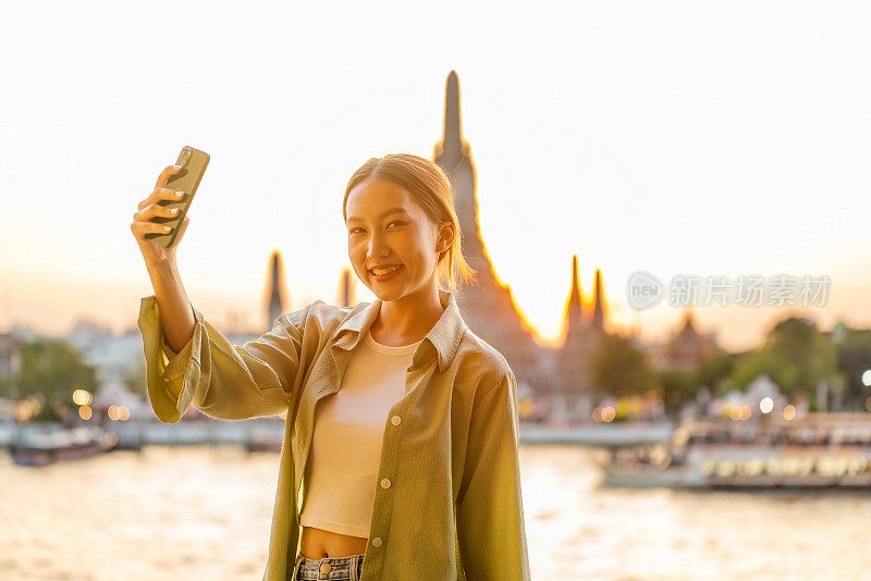 年轻的亚洲女子在泰国曼谷河边的阿龙寺的背景下捕捉自己日落的时刻