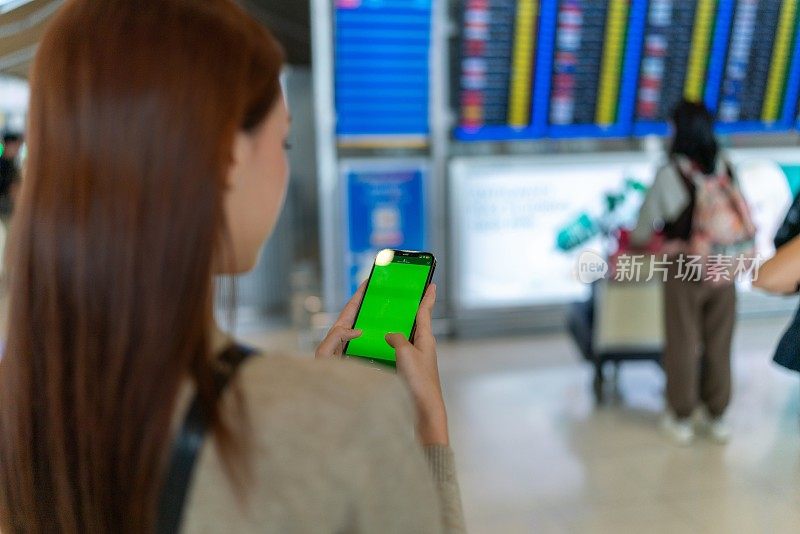 后视图:年轻的亚洲女子手持手提包，手持智能手机，站在机场候机楼的FIDS登机牌前。出差的商务人士。手持智能手机的年轻亚洲女性旅客在机场候机楼。