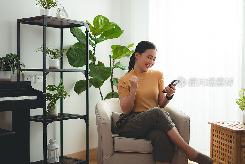 亚洲女性坐在家里的扶手椅上，用智能手机开心兴奋地做出了获胜的手势。