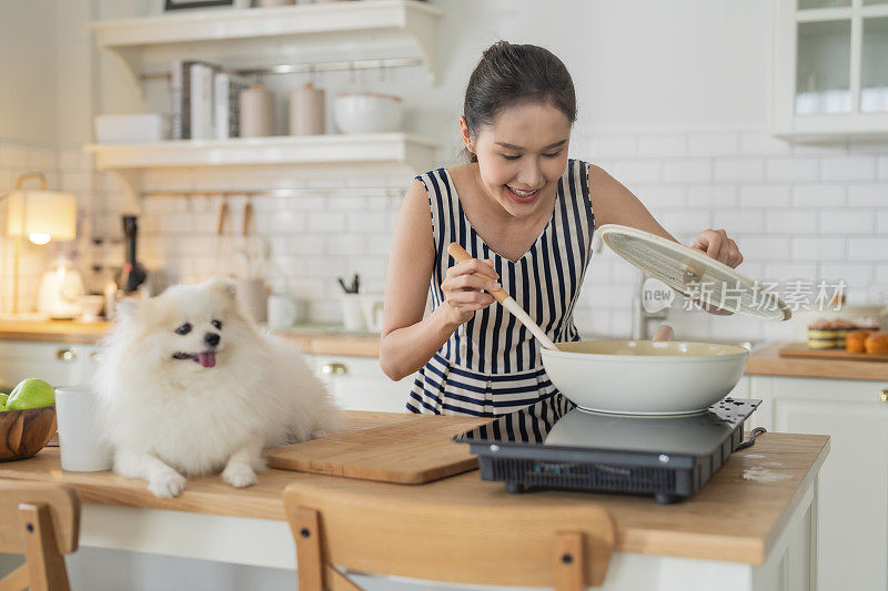 新鲜微笑的亚洲青少年女性烹饪会议与她的波美拉尼犬白色膝上狗在厨房与幸福好氛围的女人烹饪，而宠物她的狗在厨房家庭内部