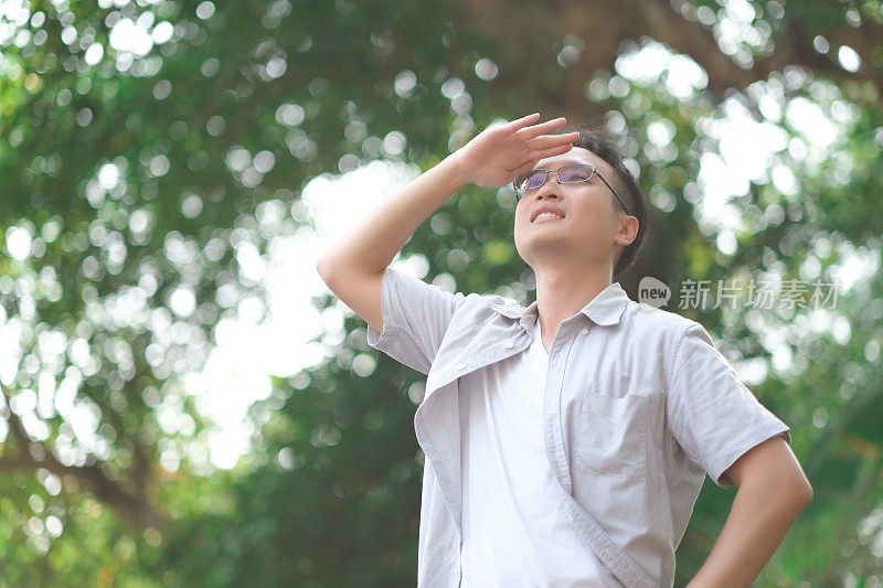 年轻的亚洲人用手遮挡阳光，抬头看周围的大自然，绿色的散景和光线闪耀着淡淡的微笑，感觉清新，人们对自然的责任观念。