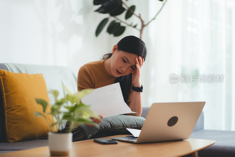 亚洲女性坐在沙发上，担心银行票据、抵押贷款债务和生活成本的账单支付问题。